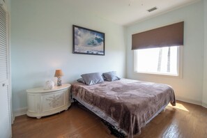 A bedroom with a bed, a nightstand, a lamp, a painting, and a window with blinds. - Here Awhile-30 Day Oceanfront Escape (Folly Beach)