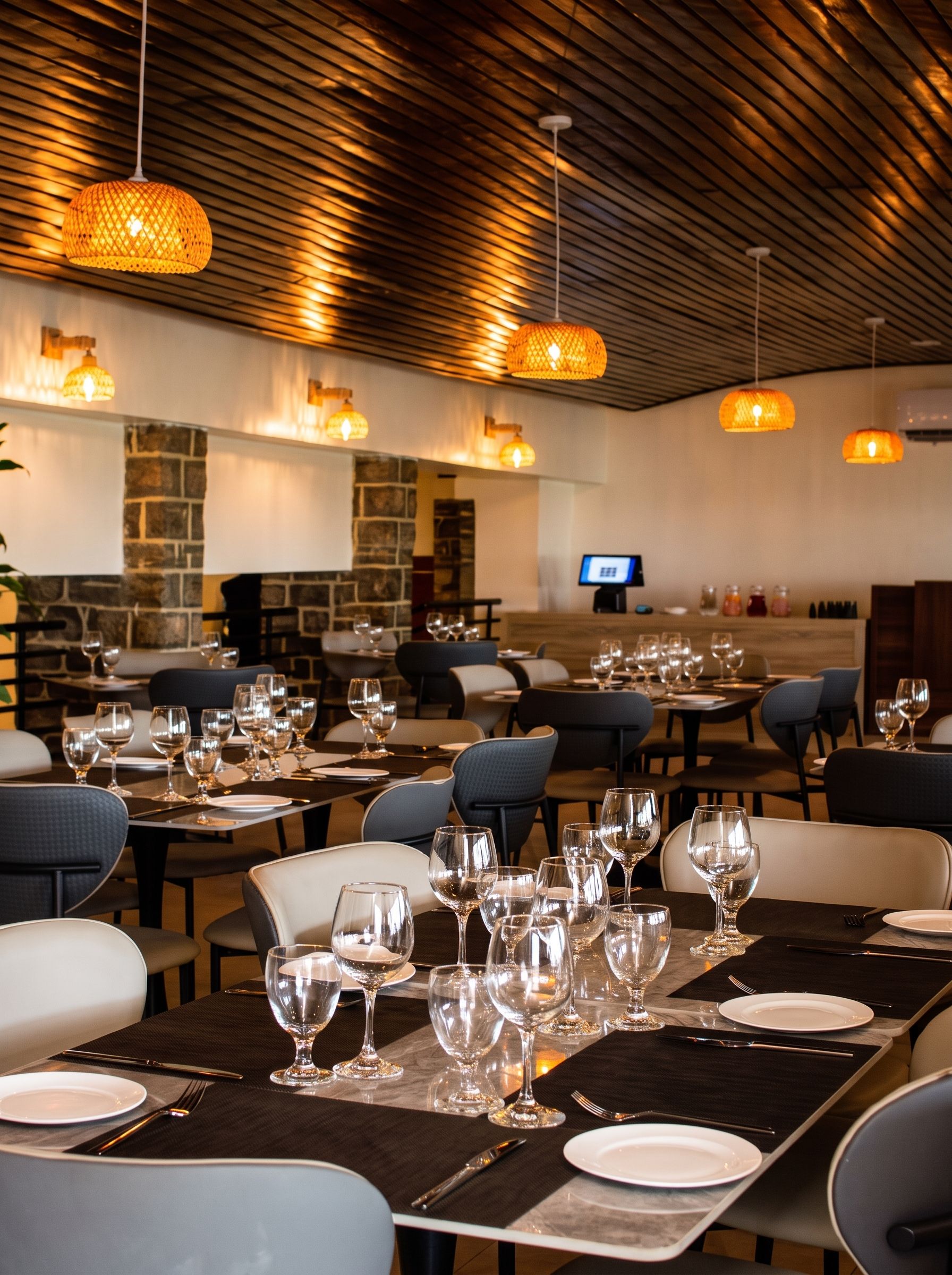 A restaurant interior with tables set for dining, a stone wall, and hanging pendant lights.