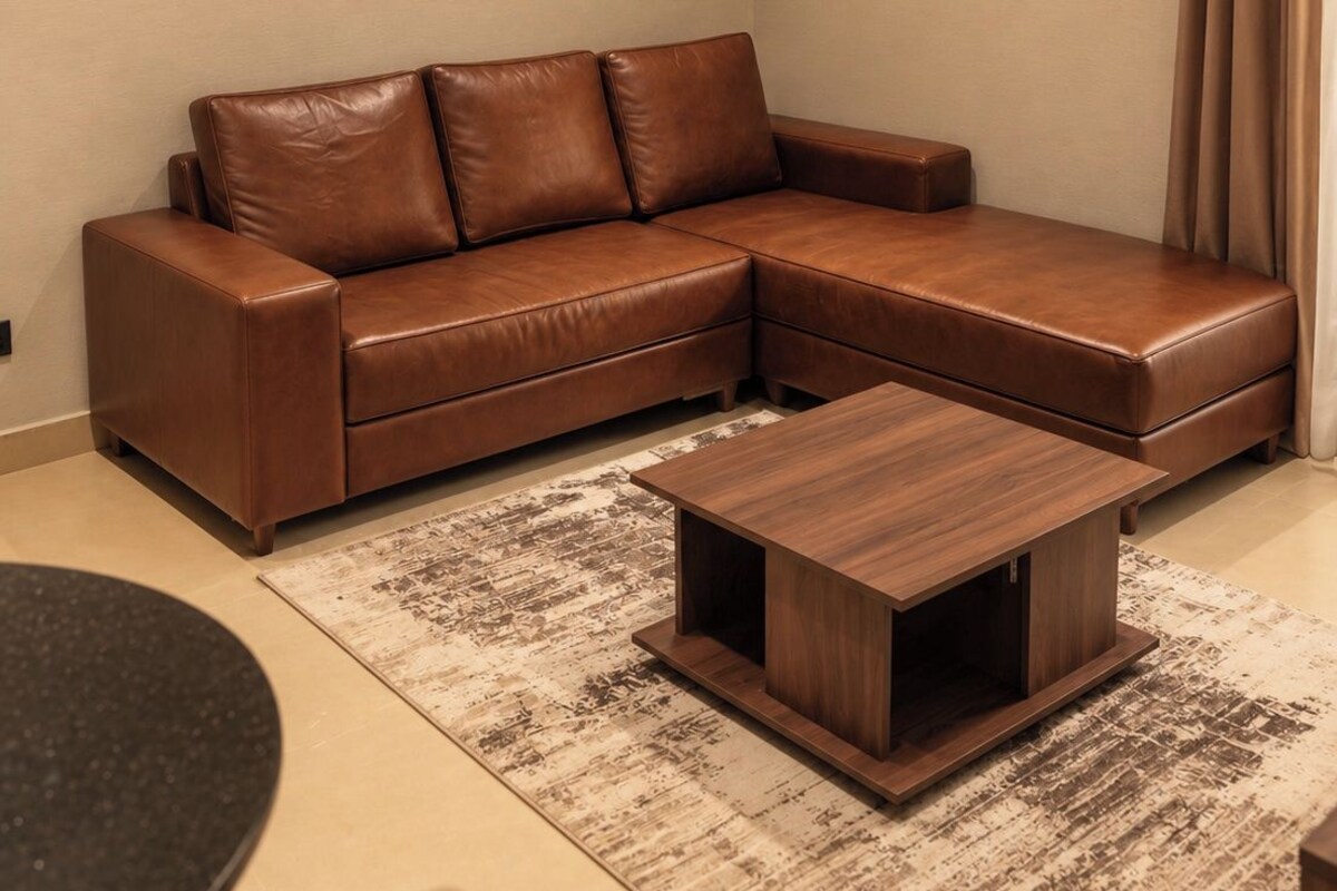 Una sala de estar con un sofá seccional de cuero marrón, una mesa de centro de madera, un cuadro enmarcado en la pared y un equipo de aire acondicionado.