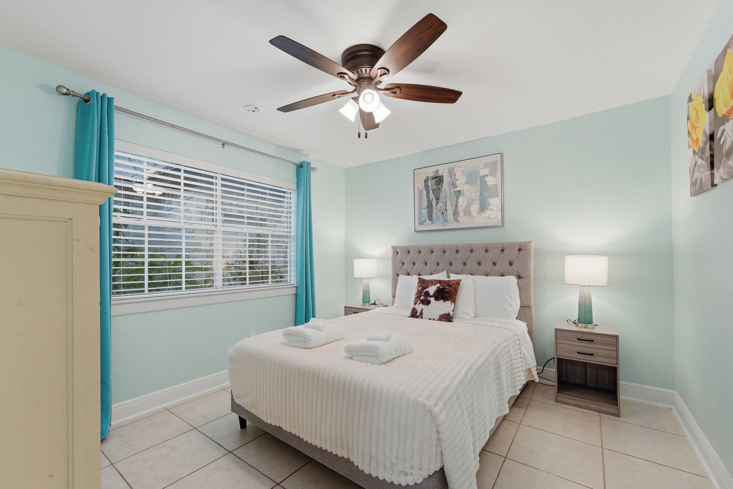A bedroom with a bed, two bedside tables, a ceiling fan, a window with blinds, and a wall-mounted artwork.