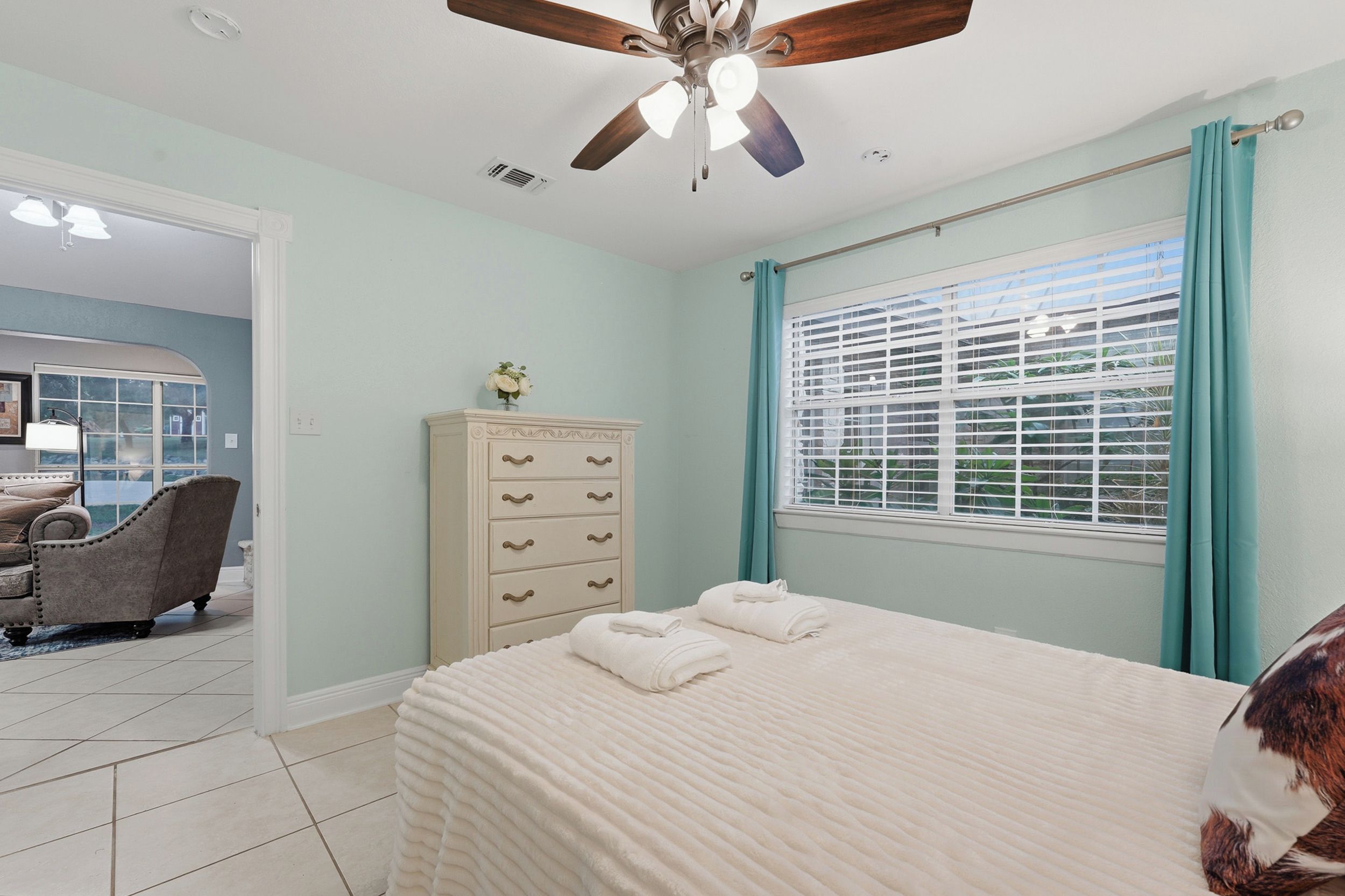 Une chambre à coucher comprenant un lit, une commode, un ventilateur de plafond et une fenêtre avec des stores.