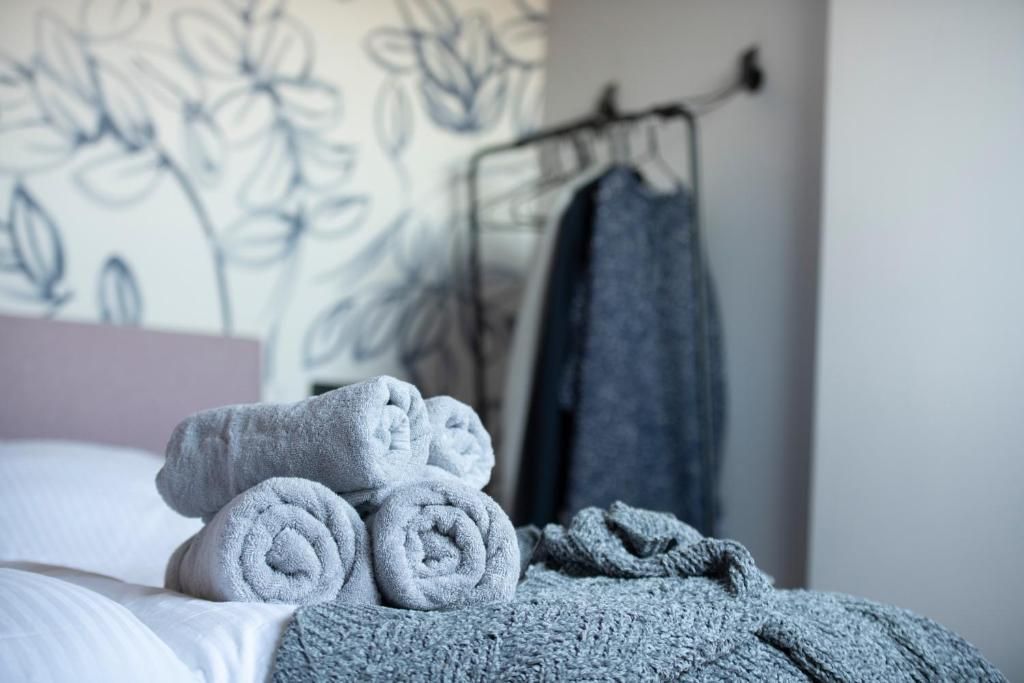 Ein ordentlich bezogenes Bett mit aufgerollten Handtüchern und einer Decke, eine Wand mit einem Blumenmuster und ein Kleiderständer mit einem Kleidungsstück.