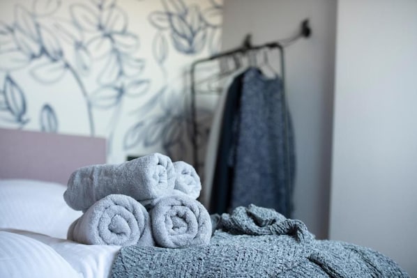 A neatly made bed with rolled towels and a blanket, a wall with a floral pattern, and a clothing rack with a garment.