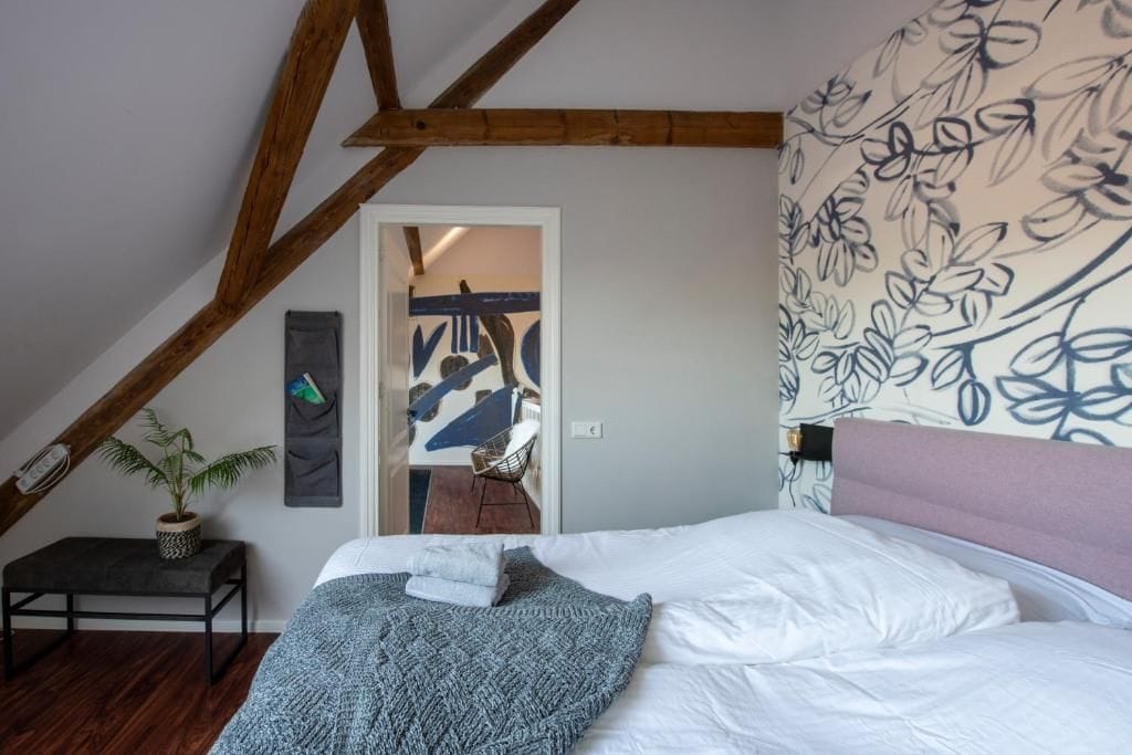 Un dormitorio con techo inclinado, una cama con ropa de cama blanca, un plaid de punto gris, una mesita con una planta y una pared con estampado floral.