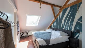 Un dormitorio con techo inclinado, una cama con ropa de cama blanca, una mesita de noche pequeña y una ventana con luz natural.