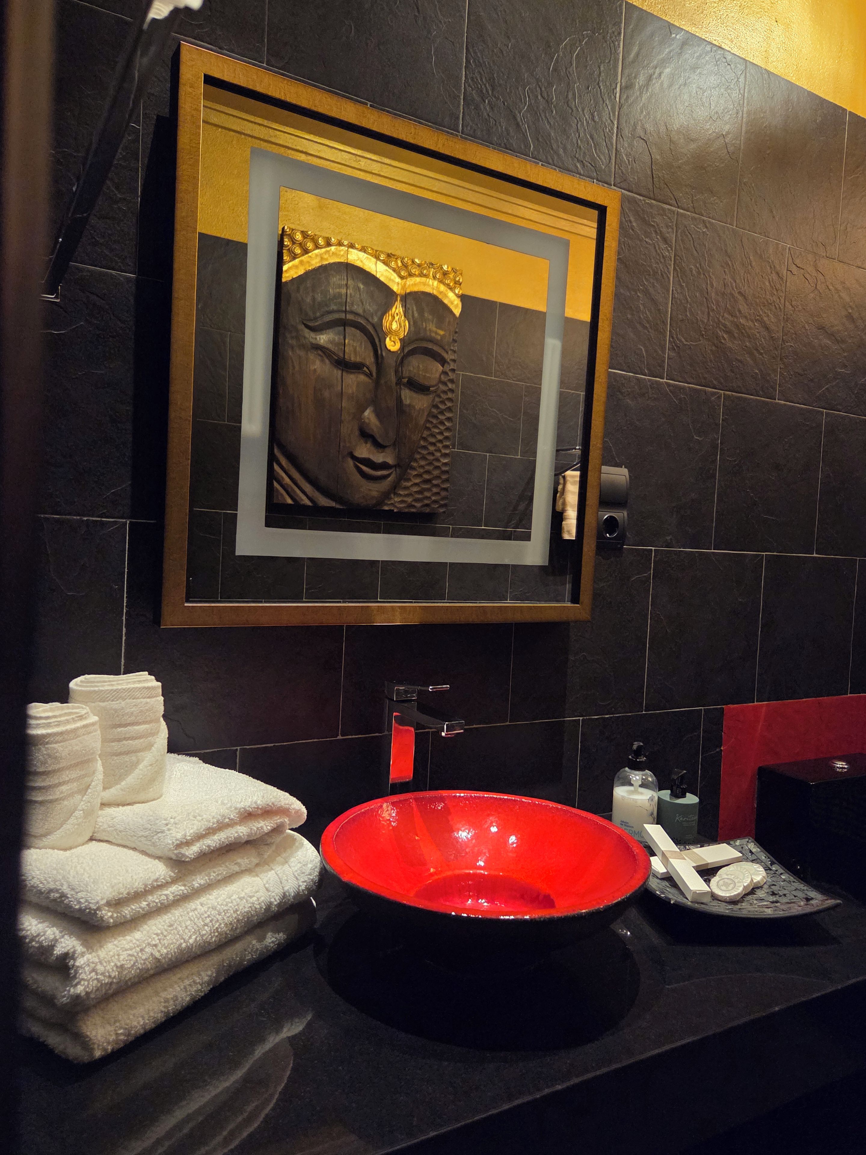 Un baño con pared de azulejos negros, una estatua de Buda enmarcada, un lavamanos rojo y toallas dobladas con cuidado.