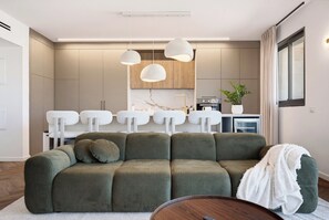 Een moderne woonkamer met een groene bank, een salontafel en een eethoek met witte stoelen en hanglampen.