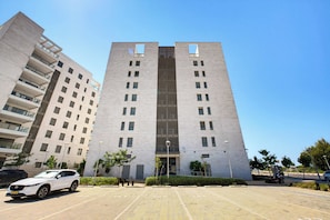 A modern multi-story building with a central entrance, surrounded by a parking area and greenery. - Premium 6BR Coastal Stay in Achziv by SeaNRent (Nahariya)