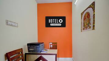 Habitación de hotel con pared de acento naranja, un escritorio con impresora, una silla y un cartel que indica que la habitación cuenta con servicio de la empresa.