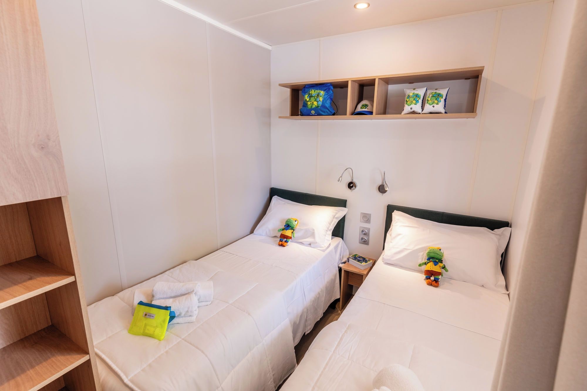 A hotel room with two beds, a shelf with items, and a wooden wardrobe.