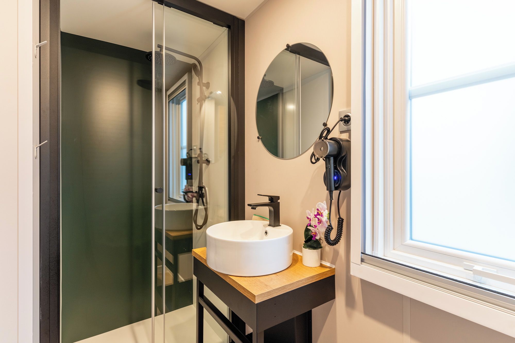 A bathroom with a white sink, a round mirror, a handheld showerhead, and a window with blinds.