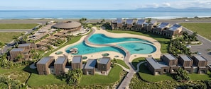 A resort with a central pool, connected by walkways, surrounded by wooden buildings, and overlooking the ocean.