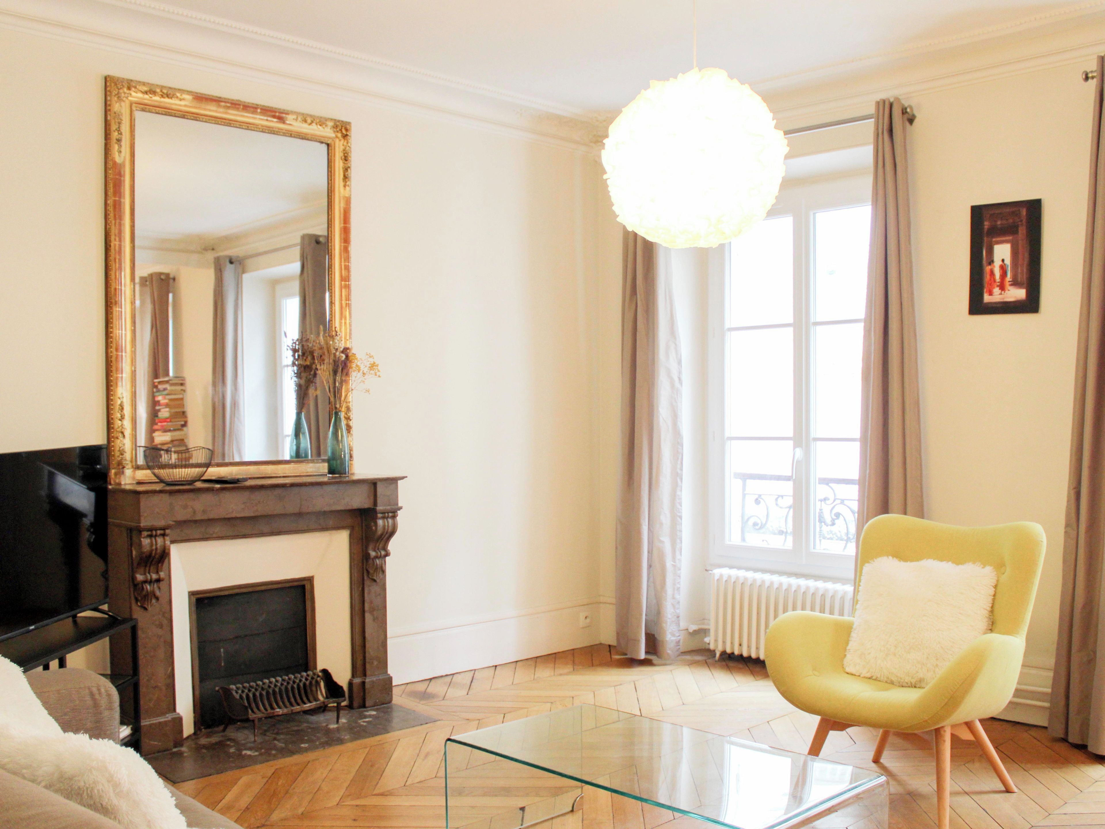 A living room with a fireplace, a large mirror, a yellow armchair, and a glass coffee table.