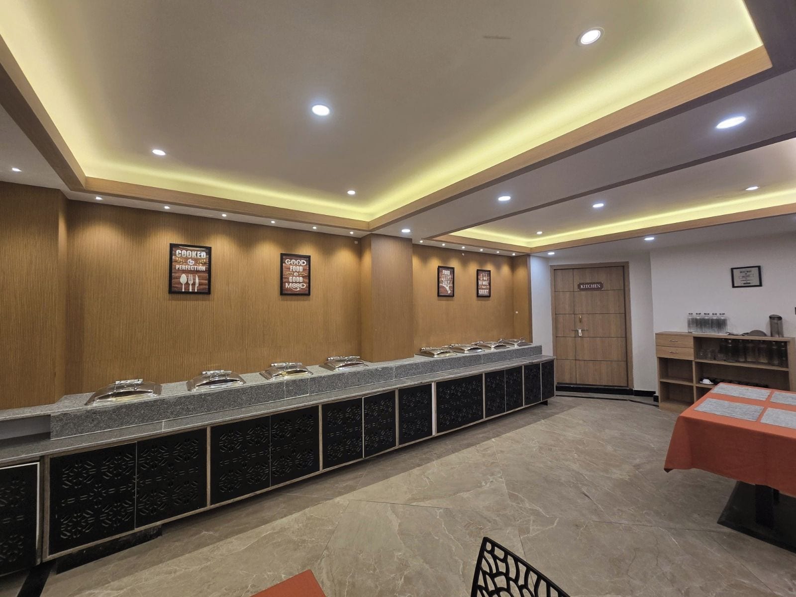 A long buffet counter with multiple serving stations, a wooden wall with framed signs, a kitchen area, and a table with a red tablecloth.
