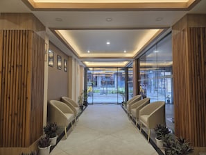 Un área de recepción con diseño moderno, que incluye una zona de estar central con sillas y plantas en macetas, y un puerta de cristal que da al exterior.