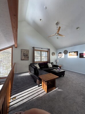 Ein geräumiges Wohnzimmer mit hoher Decke, Deckenventilator, großem Fenster, Ecksofa, Couchtisch und Kamin.