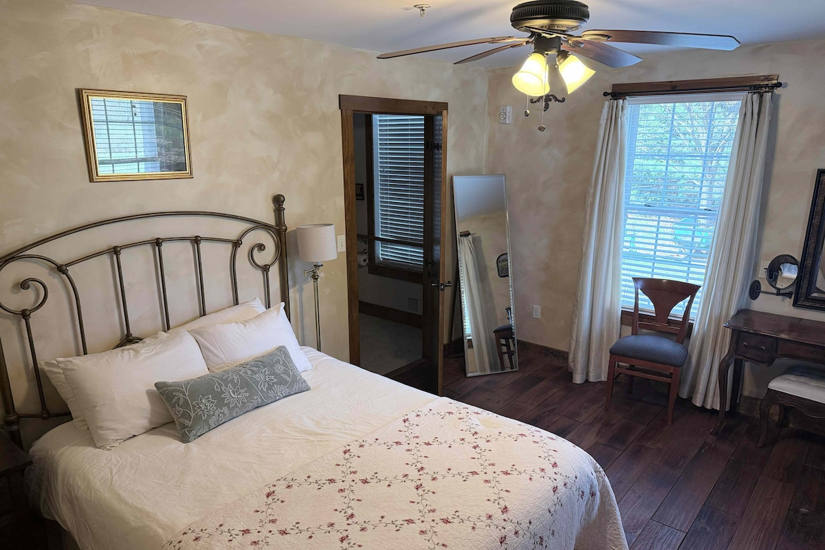 Ein Schlafzimmer mit Bett, Deckenventilator, Spiegel, Stuhl und Fenster mit Vorhängen.