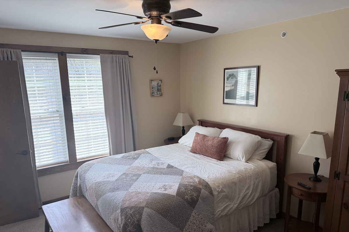 Ein Schlafzimmer mit einem Bett, einem Deckenventilator, einem Fenster mit Vorhängen, einem Nachttisch mit Lampe und einem Wandbild.