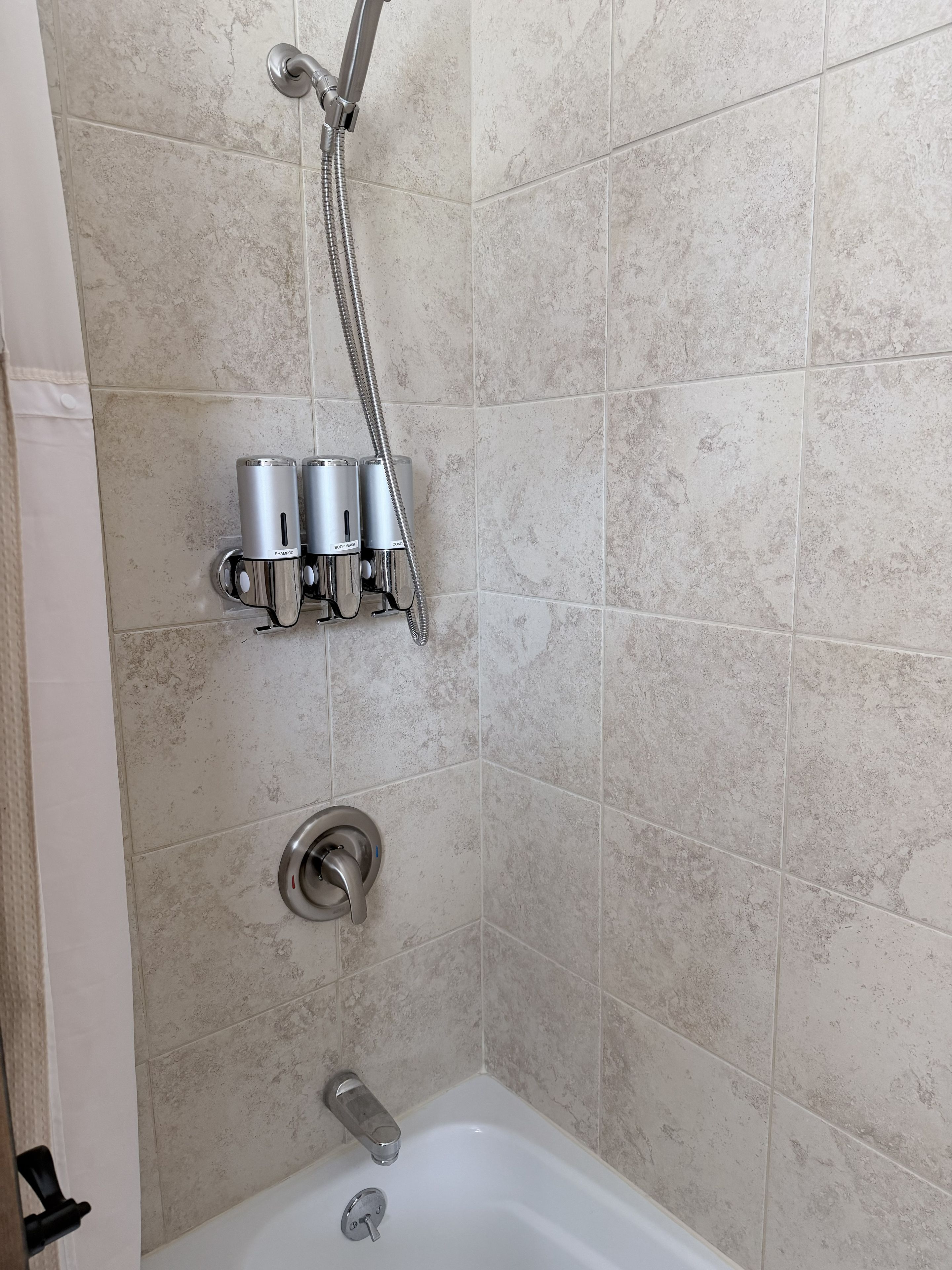 Un área de ducha con cabezal de ducha manual, grifo empotrado y dos dispensadores de jabón.