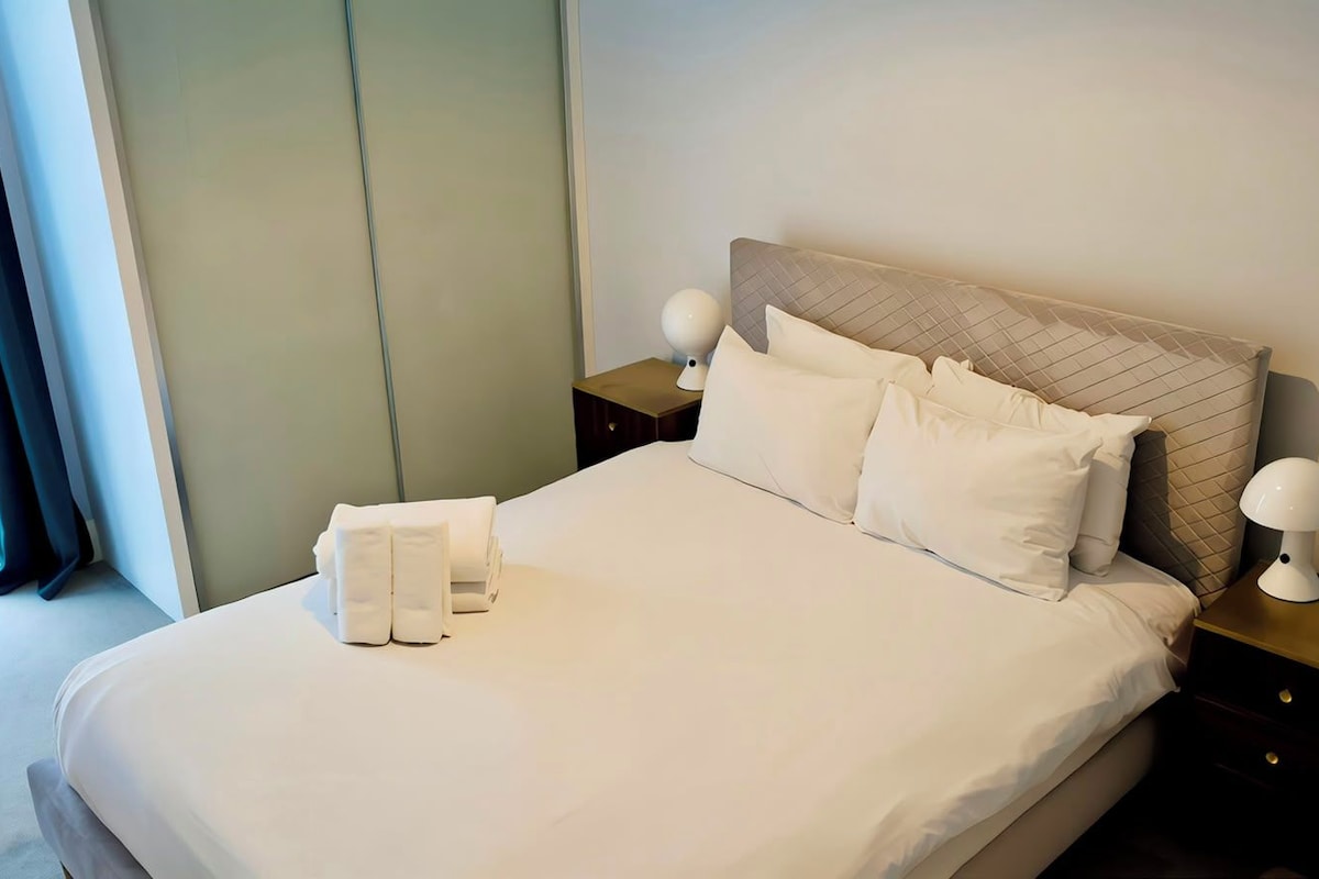 A neatly made bed with white linens, a headboard with a quilted pattern, two bedside lamps, and a folded towel on the bed.