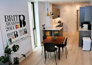 A modern kitchen with white cabinets, a dining area with a wooden table and black chairs, and a large poster on the wall advertising the BRIT Awards 2014. - The Warwick Lane Residences by Aureva (London)