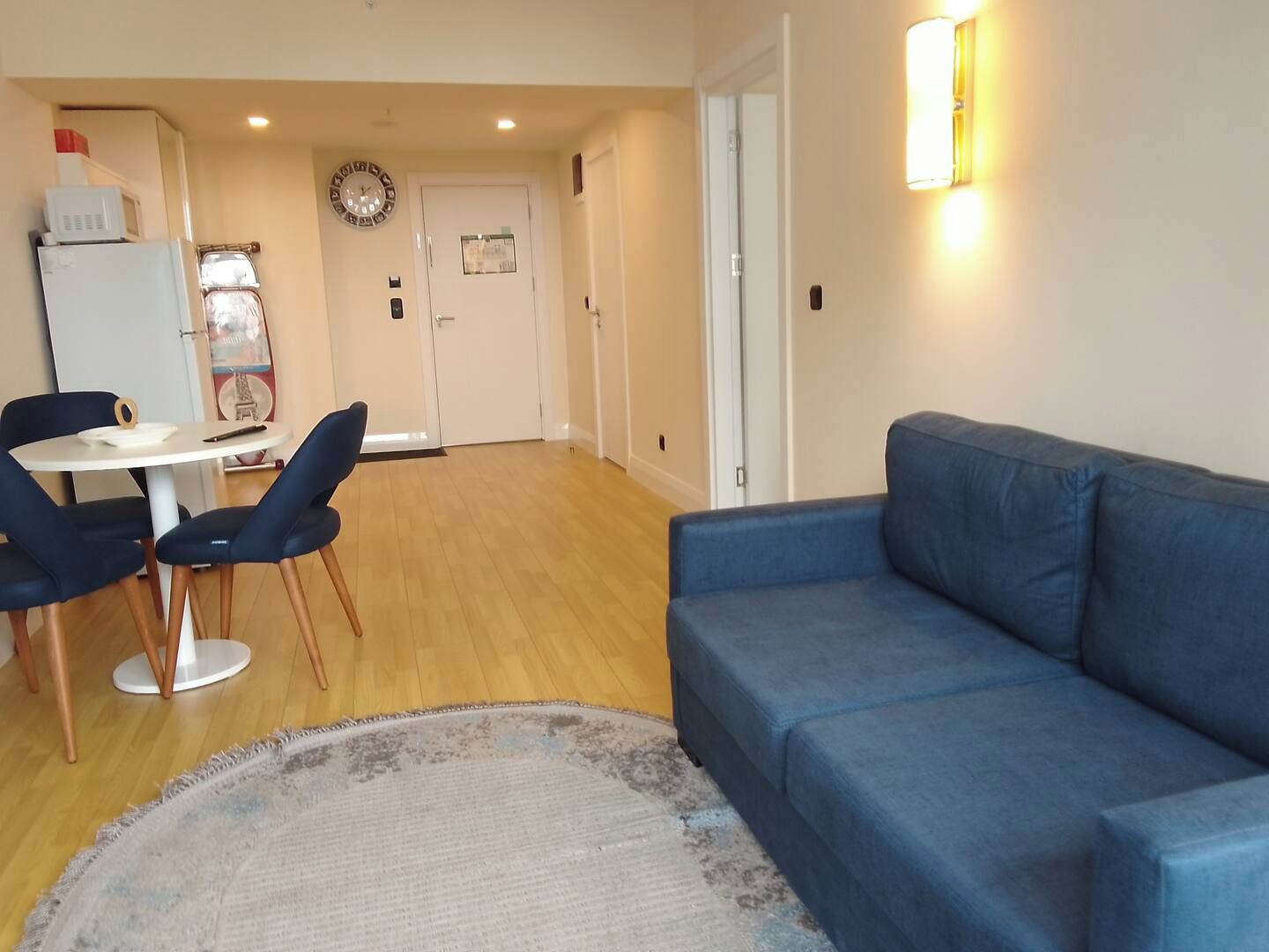 Un salon avec un canapé bleu, une table basse ronde et deux fauteuils bleus. On aperçoit une porte blanche au fond, ainsi qu’un luminaire fixé au mur.