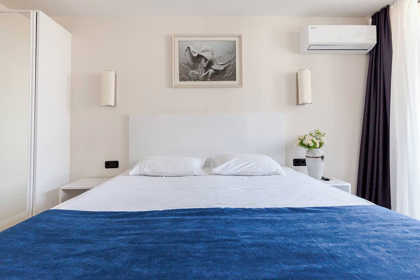Une chambre d’hôtel comprenant un lit, une table de chevet, un climatiseur fixé au mur, un tableau encadré et un vase avec des fleurs.