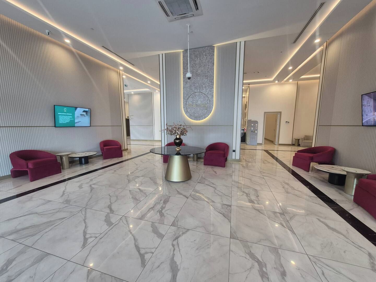 A modern lobby with a marble floor, red seating, a central table, and a mounted TV.