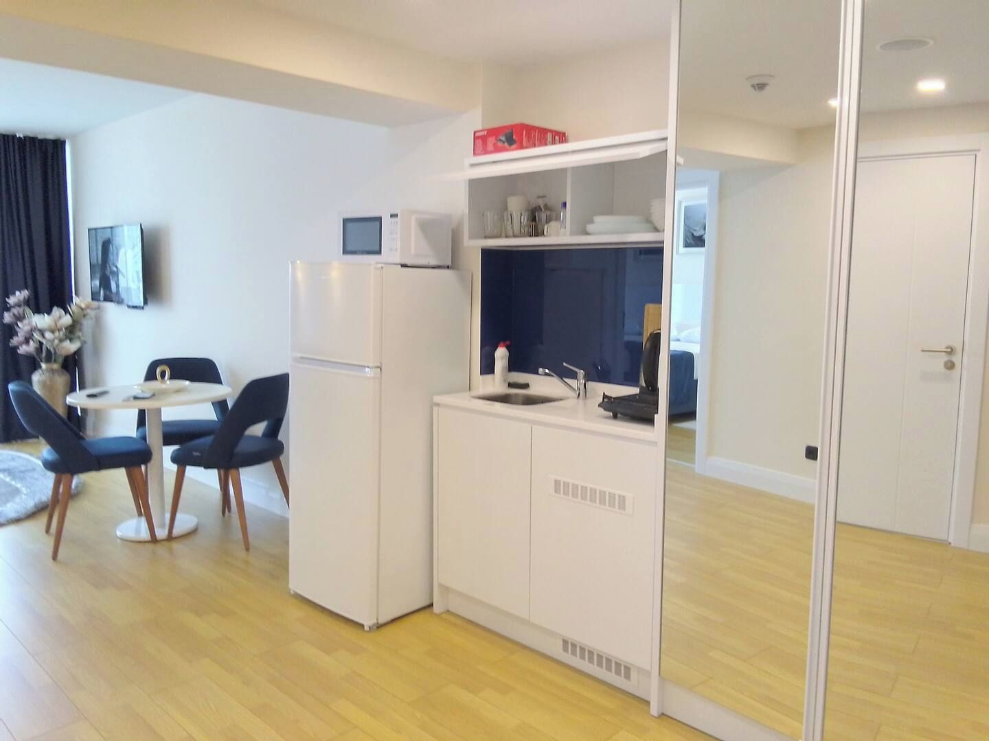 Een kleine keuken met een koelkast, magnetron en gootsteen. Er is een zithoek met twee stoelen en een tafel.