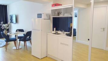 Een kleine keuken met een koelkast, magnetron en gootsteen. Er is een zithoek met twee stoelen en een tafel.
