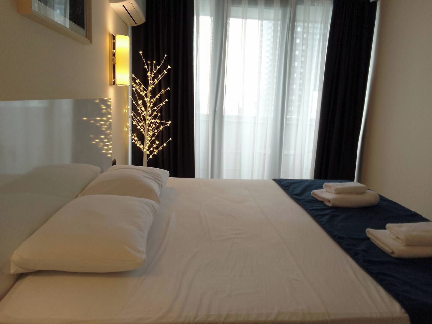 Ein Hotelzimmer mit Bett, Kissen, einem Nachttisch, einer Wandlampe und einem Fenster mit Vorhängen.