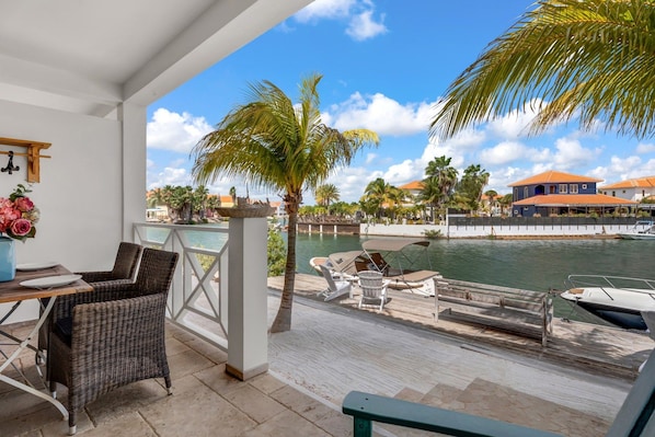 A covered outdoor area with wicker furniture, a table with plates, a vase with flowers, and a view of a body of water with boats and buildings in the background. - Ground-Floor 1BR, Marina View, Sleeps 3 · Apt 22 Ocean Breeze (Kralendijk)