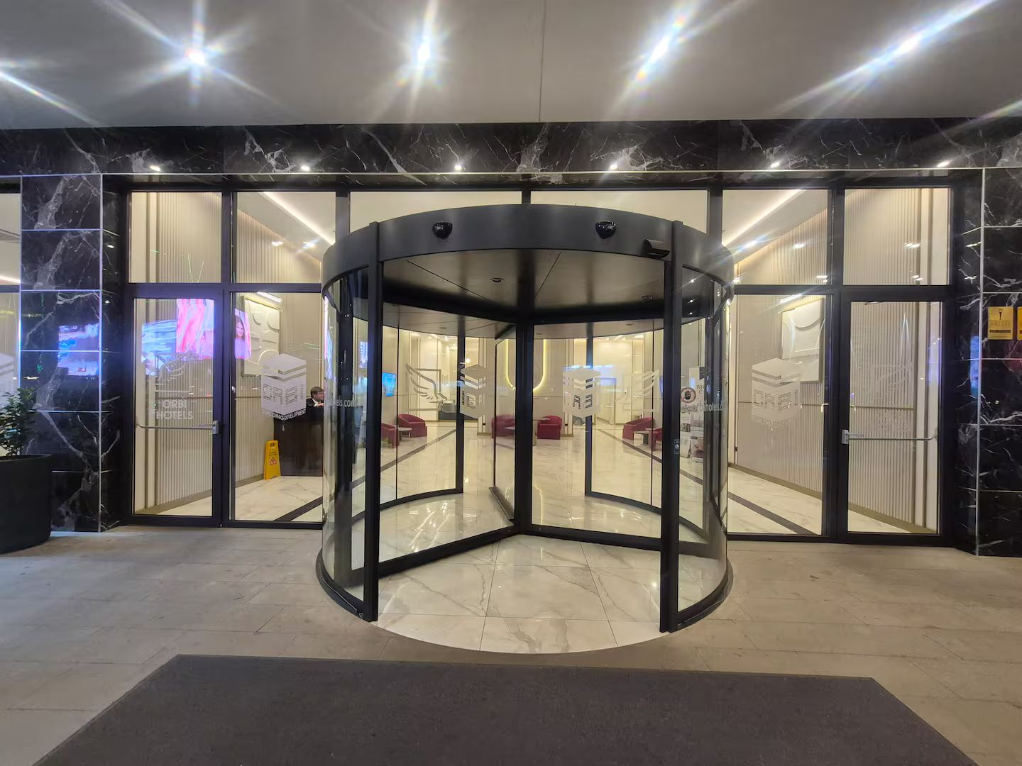 Entrada de un hotel con puerta giratoria, paredes de vidrio y mostrador de recepción.