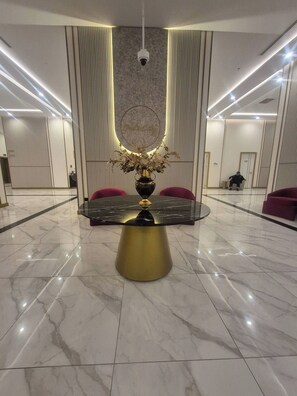 Un vestíbulo de hotel moderno con piso de mármol, una mesa redonda central con base dorada y sillones tapizados en rojo.