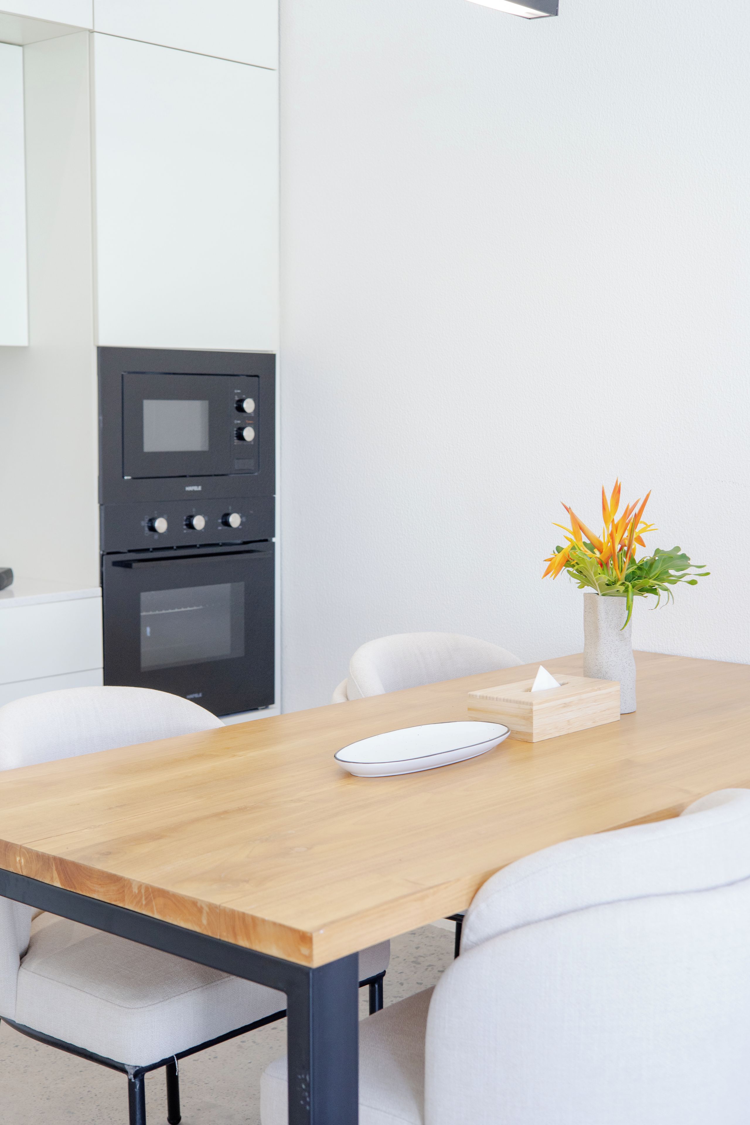 A modern kitchen with a wooden table, white chairs, a white vase with flowers, and a built-in oven.
