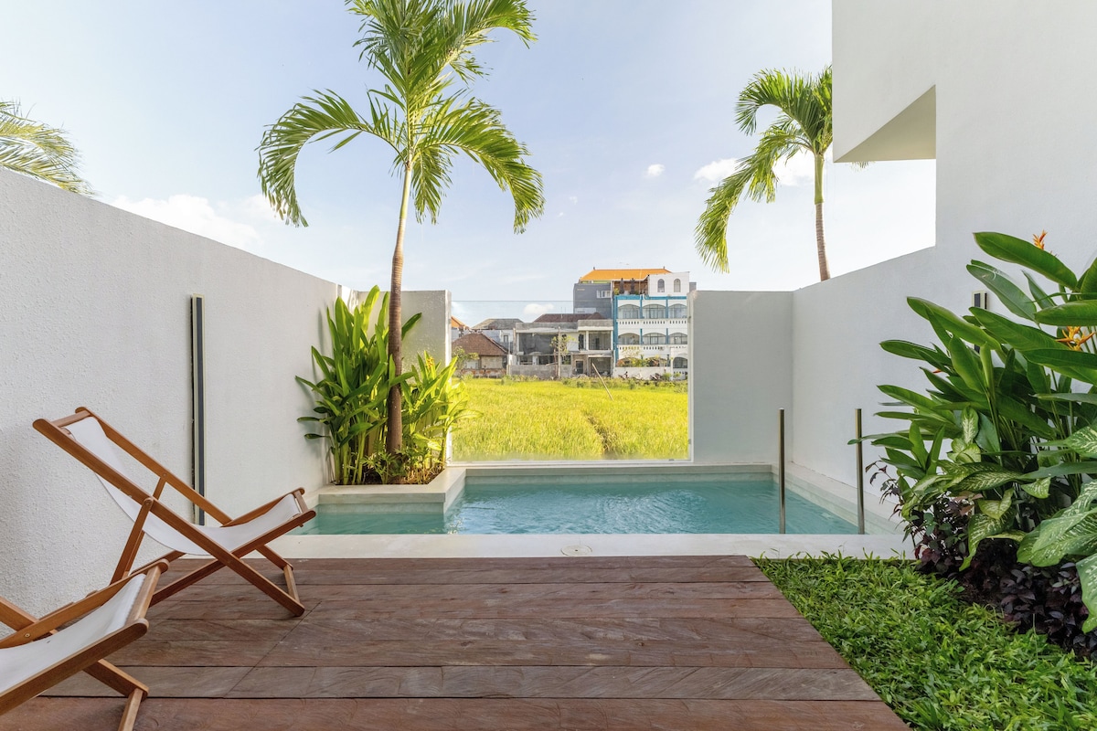 A pool area with wooden deck, lounge chairs, and a view of a green field and buildings.