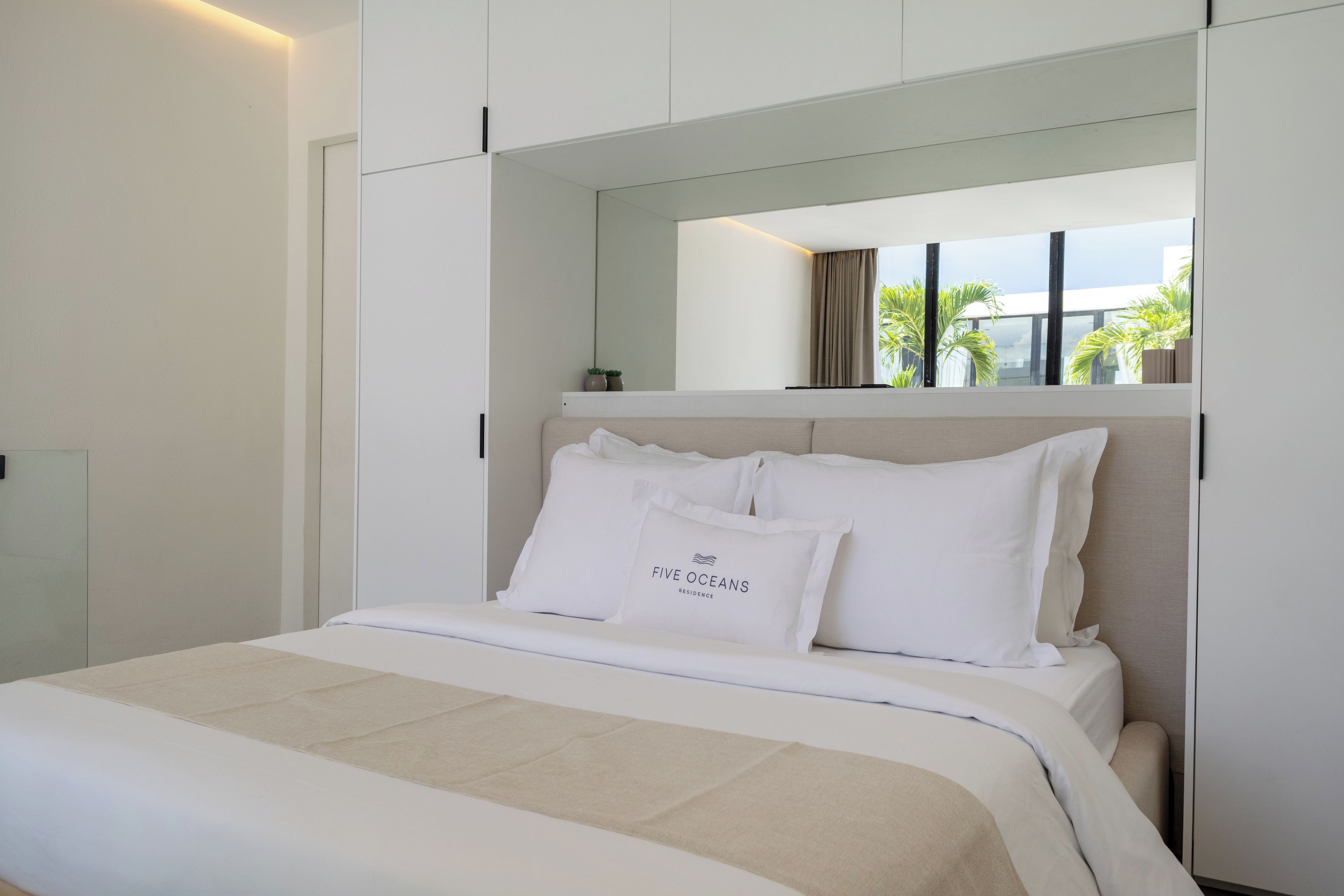 A hotel room with a bed, a window with a view of palm trees, and a sign reading 'FIVE OCEANS RESIDENCE'.
