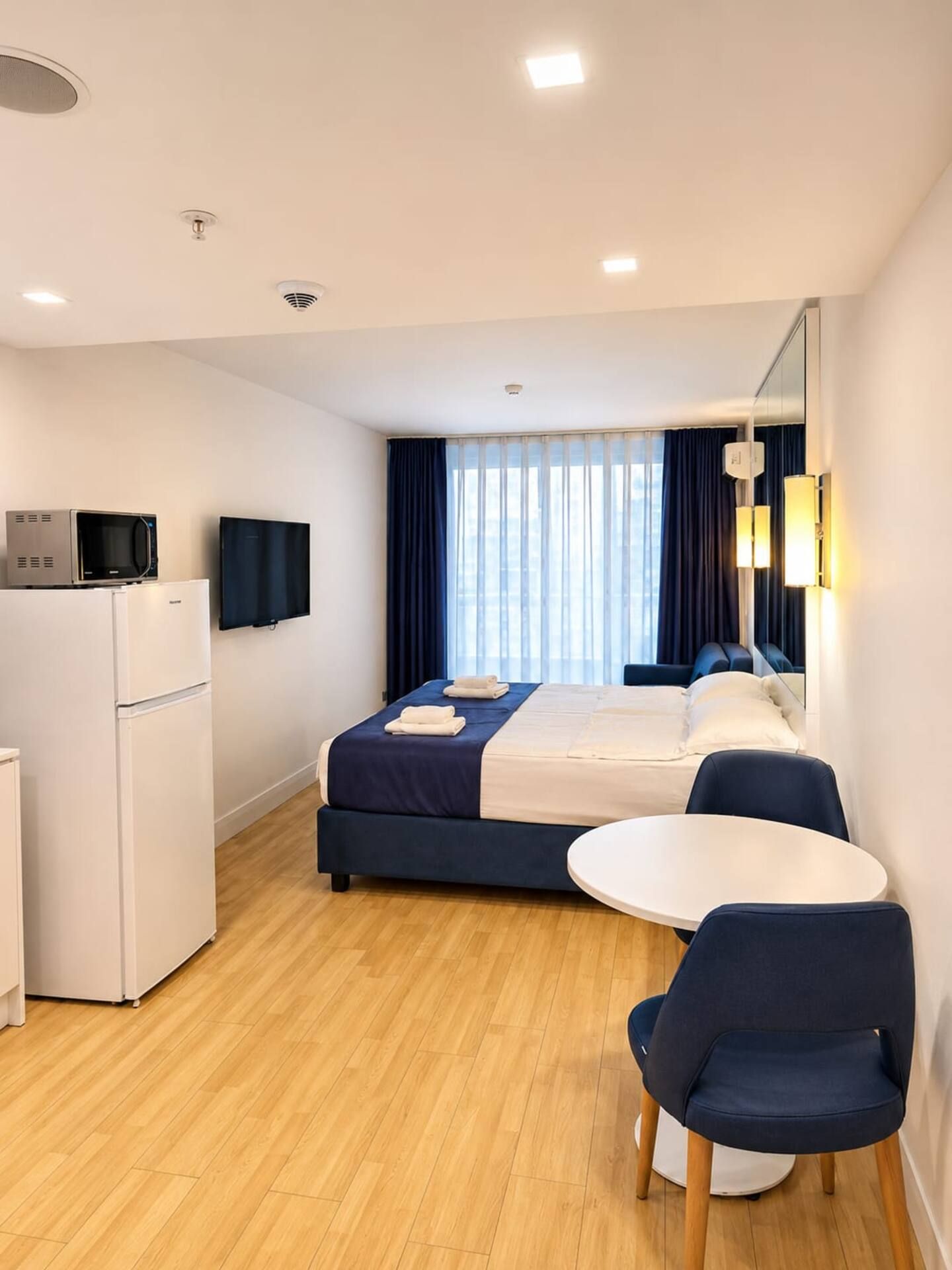 A hotel room with a bed, a desk, a chair, a microwave, a TV, and a window with curtains.