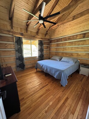 Ein Schlafzimmer einer Holzhütte mit einem Bett, Vorhängen, einem Deckenventilator und Holzboden.