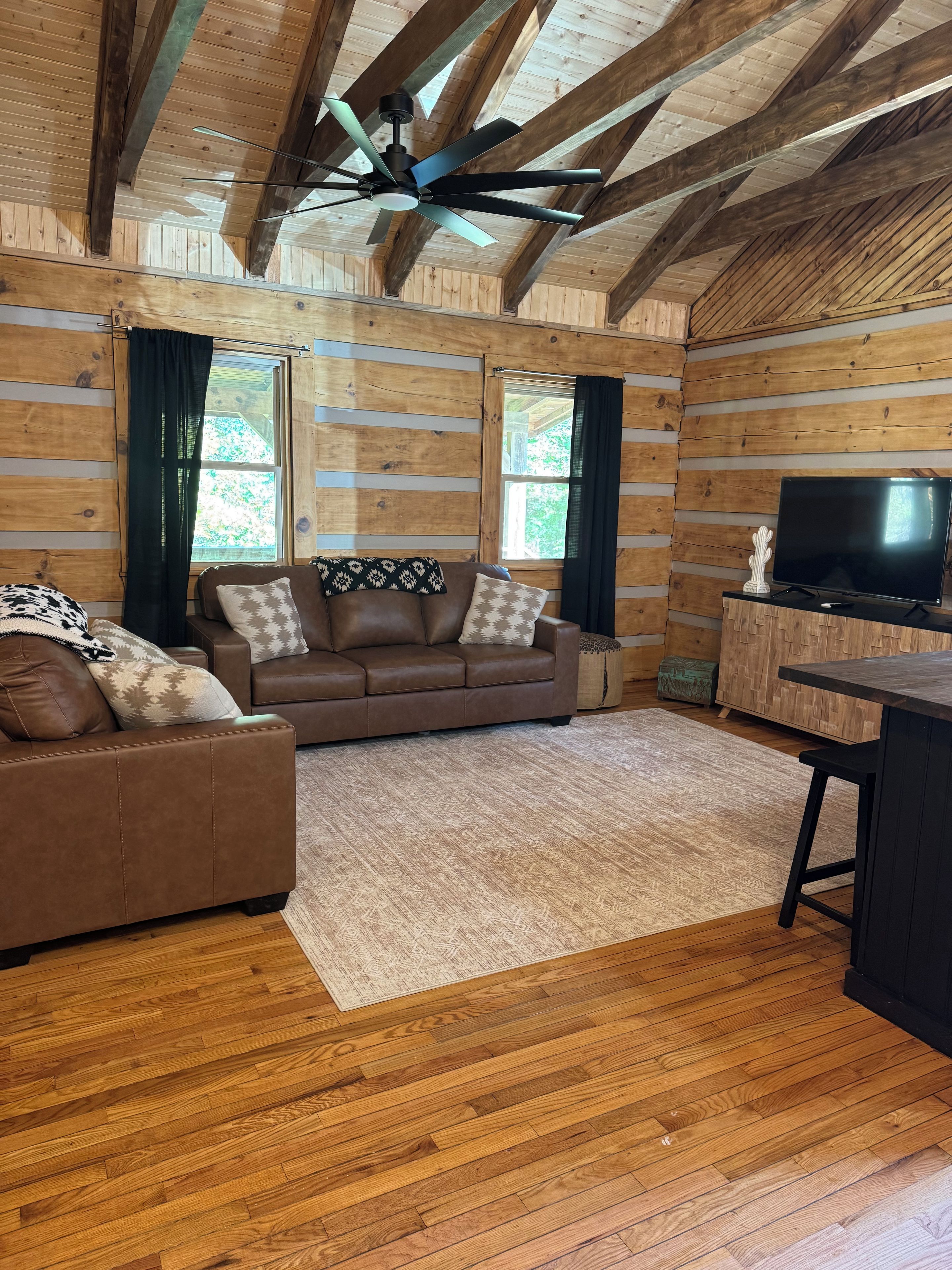 A log cabin interior with a brown leather sofa, a television on a wooden cabinet, and a ceiling fan.