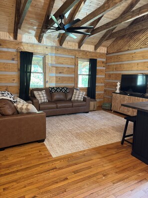 A log cabin interior with a brown leather sofa, a television on a wooden cabinet, and a ceiling fan.
