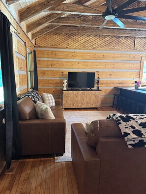 A cozy living room with wooden walls and ceiling, a brown leather sofa, a television, and a dining area with a table and chairs.