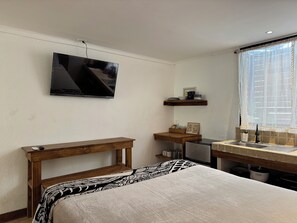 A bedroom with a bed, a bedside table, a TV mounted on the wall, a window with curtains, and a kitchen area with a sink and countertop. - New Listing (Santa Teresa)