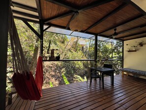 A wooden deck with a table and chairs, a hanging hammock, and a view of a lush garden through large windows. - New Listing (Santa Teresa)