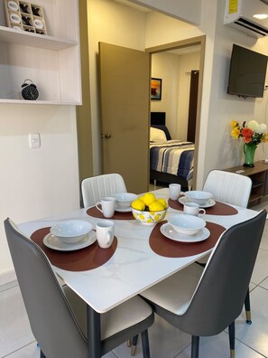 Une chambre d’hôtel avec une table à manger dressée pour quatre personnes, un lit recouvert d’une courtepointe à rayures, un téléviseur fixé au mur et une étagère ornée d’objets décoratifs.