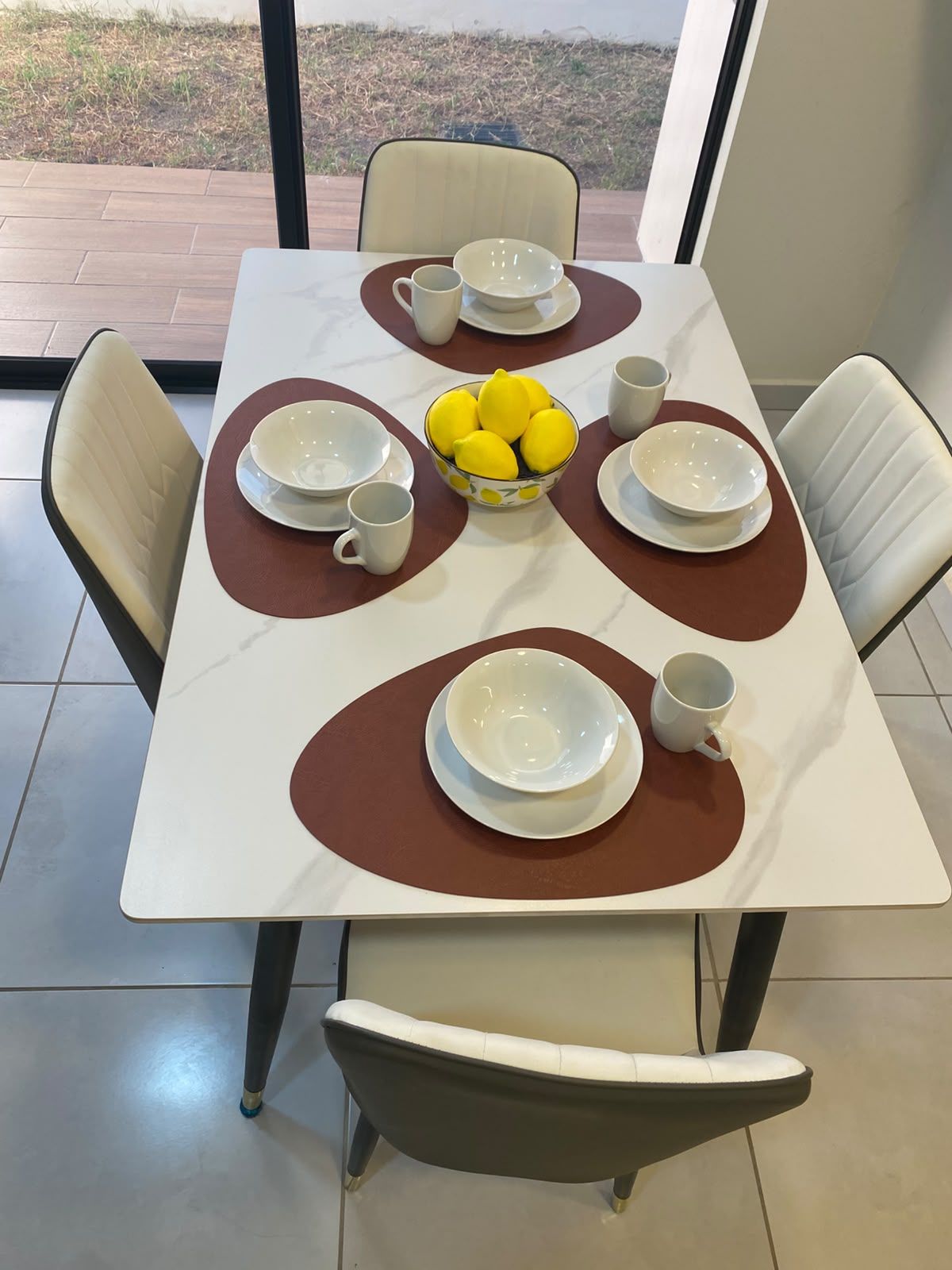 Une table à manger avec des assiettes et des bols blancs, ainsi qu’un saladier rempli de citrons. Six chaises sont disposées autour de la table.