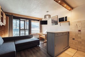 A compact living space with a sofa, a dining table, a kitchen area, and a view through a large window. - Glaciers (Les Belleville)