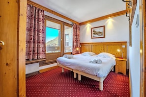 A hotel room with a bed, a nightstand, a lamp, a window with curtains, and a picture on the wall. - Glaciers (Les Belleville)