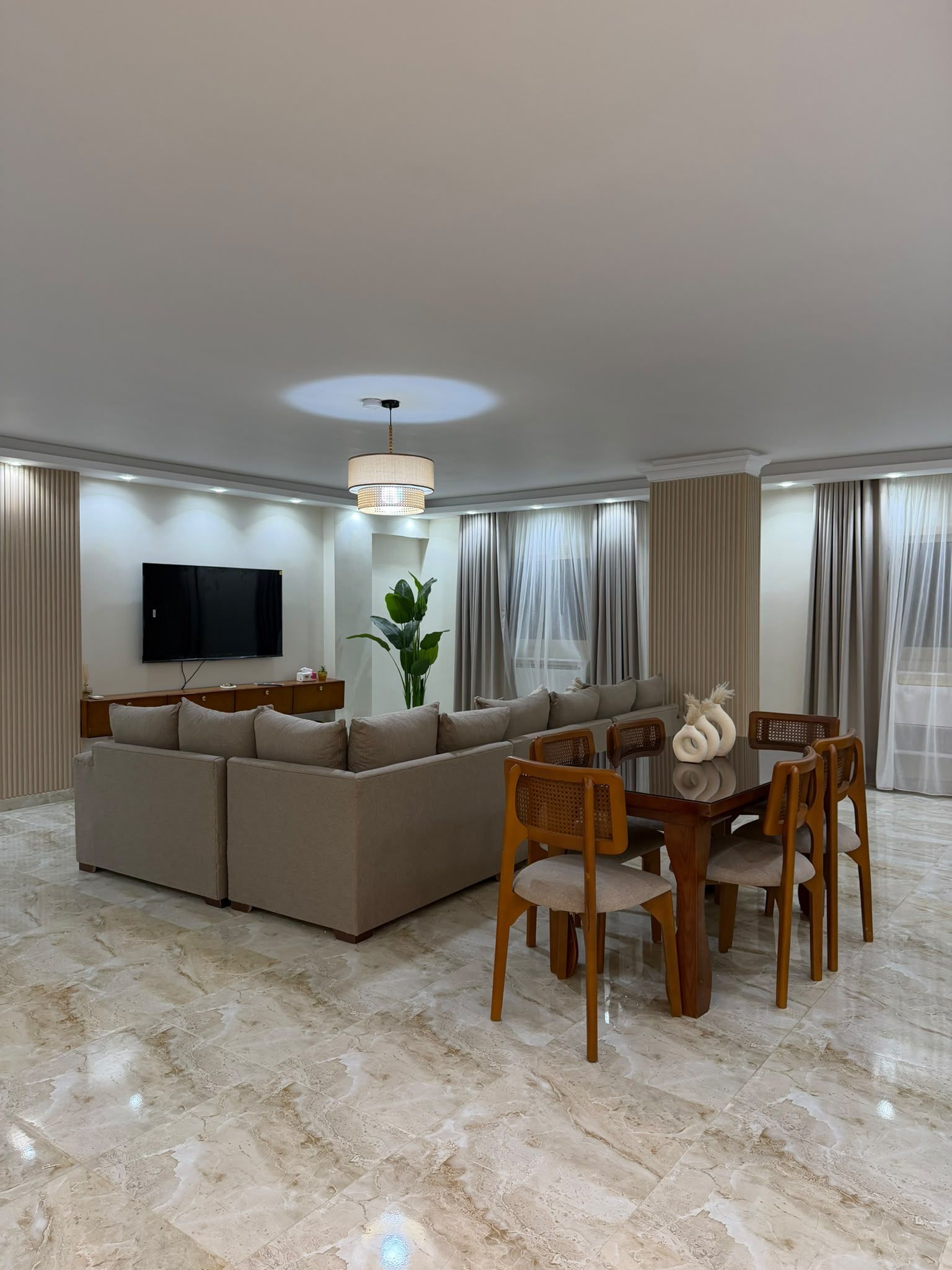 A modern living room with a sectional sofa, a dining table with chairs, a flat-screen TV mounted on the wall, and a large plant.