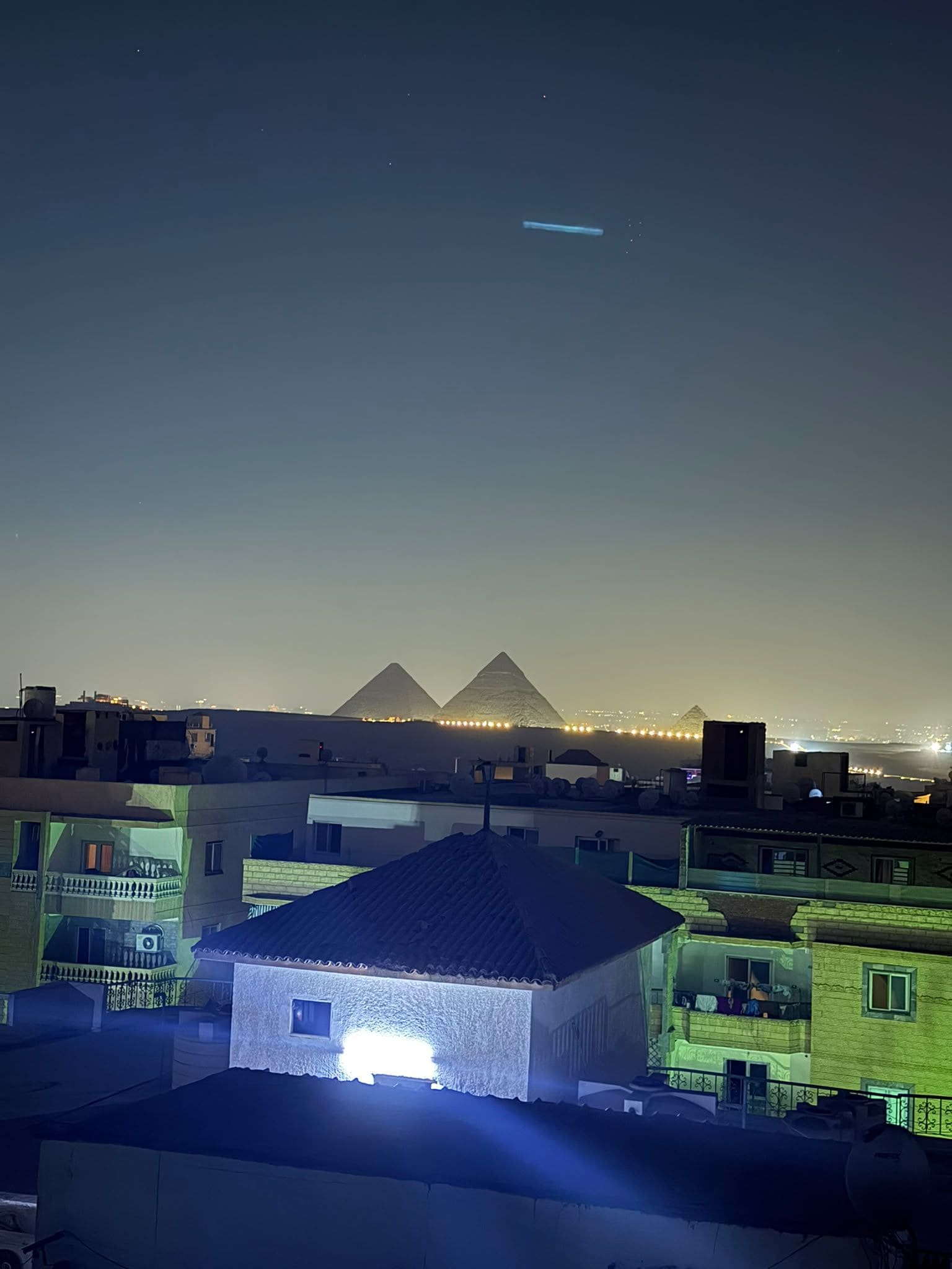 A nighttime view of a cityscape with illuminated buildings and pyramids in the distance.